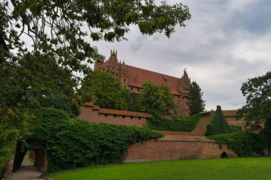 A stunning castle stands majestically amid vibrant greenery under a cloudy sky. The structure features intricate architecture and is enveloped by thick foliage and grassy grounds.