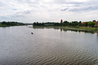 A small boat glides across a serene river reflecting the overcast sky. In the distance, a historic tower stands near lush greenery, creating a tranquil atmosphere in the landscape.