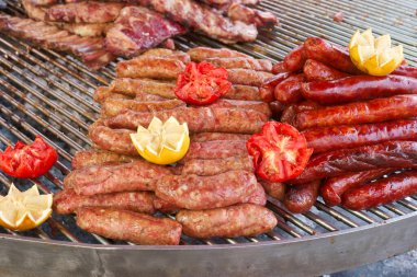 Grilled sausages and meats are arranged on a large barbecue grill, garnished with vibrant tomatoes and lemon slices. The scene captures the lively atmosphere of a summer outdoor event.