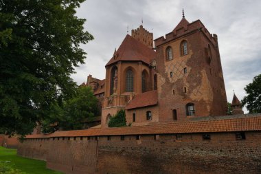 A majestic brick castle stands tall with its turrets and intricate architecture. Lush trees frame the scene, creating a serene atmosphere under a cloudy sky.