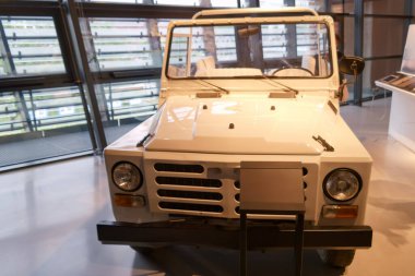 A white utility vehicle is showcased inside a contemporary museum. Large windows let in natural light, enhancing the vehicle's simple and rugged design.
