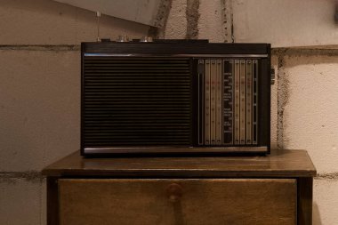A vintage radio sits on a wooden cabinet in a basement. The rough concrete walls and low light create a cozy, nostalgic atmosphere reminiscent of past decades.