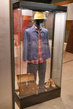 A historic mining outfit is featured in a glass case at a museum. This display includes a plaid shirt, overalls, boots, and a hard hat, illustrating mining culture.