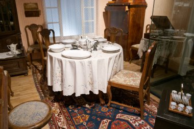 Parlak yemek odasında beyaz tabaklar ve çatal bıçaklarla dolu yuvarlak bir masa var. Antika ahşap sandalyeler masayı sarıp sıcak bir atmosfer yaratıyor. Klasik dekor cazibe katar.