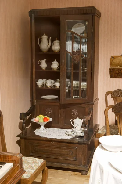 Güzel hazırlanmış ahşap bir dolap sıcak bir yemek odasında duruyor, narin porselenleri sergiliyor. Bir kase taze meyve renk ve cazibe ekler. Doğal ışık davetkar atmosferi güçlendirir.
