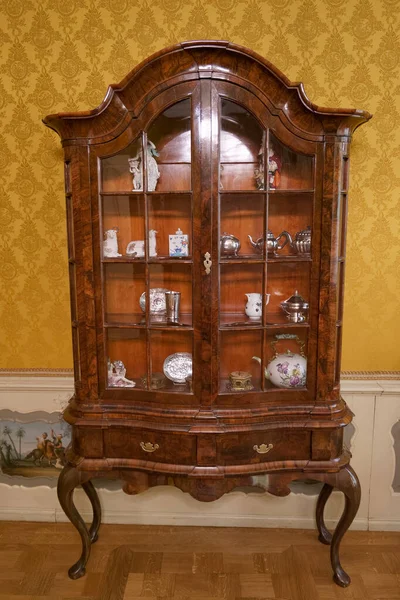 Antika bir vitrin, rahat bir odada, nefis porselenler ve çatal bıçaklarla gururla duruyor. Dolapta cam kapılar ve dekoratif detaylar var..