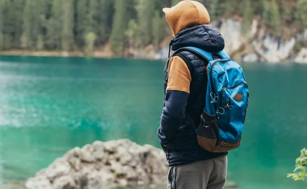 The boy walks on the mirror lake in the mountains, in a brown jacket and a blue backpack