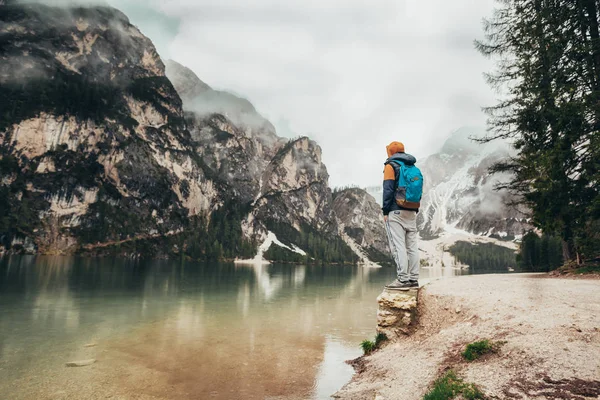 The boy walks on the mirror lake in the mountains, in a brown jacket and a blue backpack