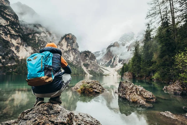 The boy walks on the mirror lake in the mountains, in a brown jacket and a blue backpack