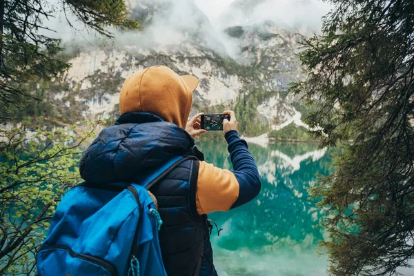 The boy walks on the mirror lake in the mountains, in a brown jacket and a blue backpack