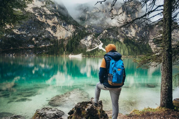 The boy walks on the mirror lake in the mountains, in a brown jacket and a blue backpack