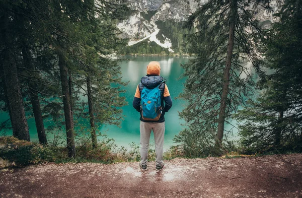 The boy walks on the mirror lake in the mountains, in a brown jacket and a blue backpack