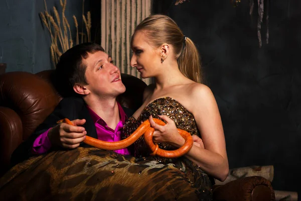 Beautiful couple in evening dress and dress suit sits with orange corn snakes, smiles and looks at each other