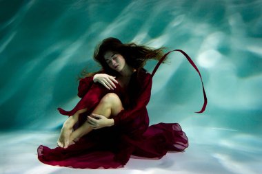 Kırmızı elbiseli bir havuzda su altında dans eden kadın.