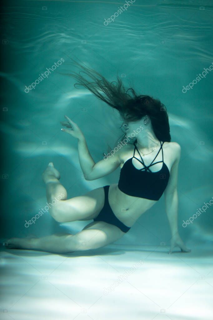 Dancing woman under the water in a pool in a black clothes.