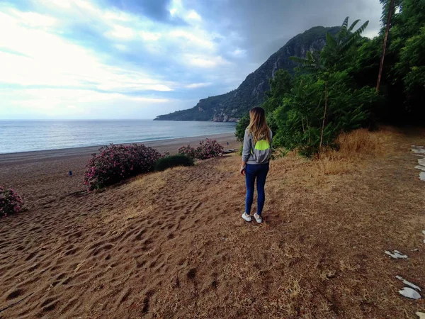 Giovane bella ragazza con la camicia con cappuccio e zaino turistico guardando sul paesaggio incredibile di montagna vicino al mare con spiaggia di pietra e cielo blu. Spiaggia Olympos, Turchia — Foto Stock