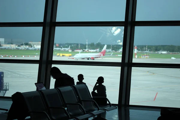 Silhouetten von Familien, die am Fenster sitzen und auf die Landebahn des Flughafens mit Flugzeugen blicken und auf ihren Flug warten — Stockfoto