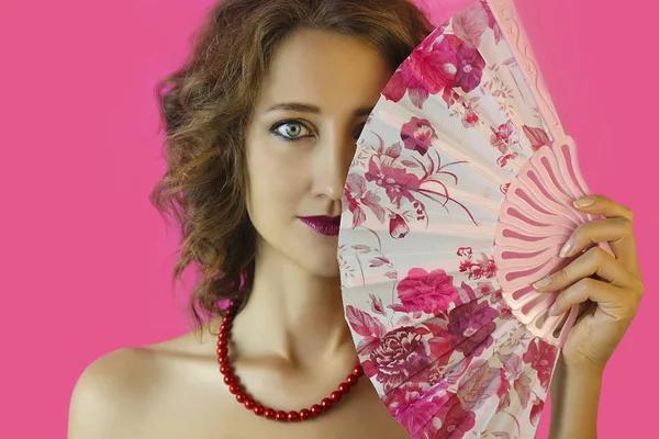 Parlak makyaj ve eller yakın çekim pembe bir arka plan üzerinde bir fan ile güzel bir genç kız portresi.