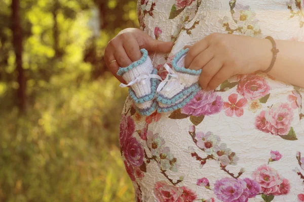 Jovem grávida. Mãos de uma menina grávida segurando um close-up de botas de bebê no parque — Fotografia de Stock