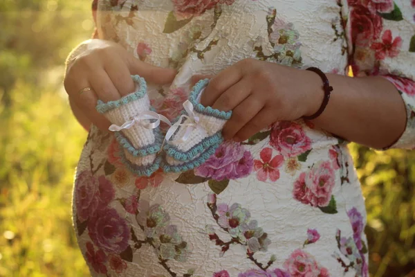 Jovem grávida. Mãos de uma menina grávida segurando um close-up de botas de bebê no parque — Fotografia de Stock