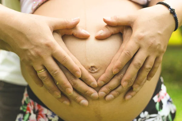 Jovem casal bonito cruzou as mãos na forma de um coração na barriga de uma menina grávida close-up na temporada de verão — Fotografia de Stock