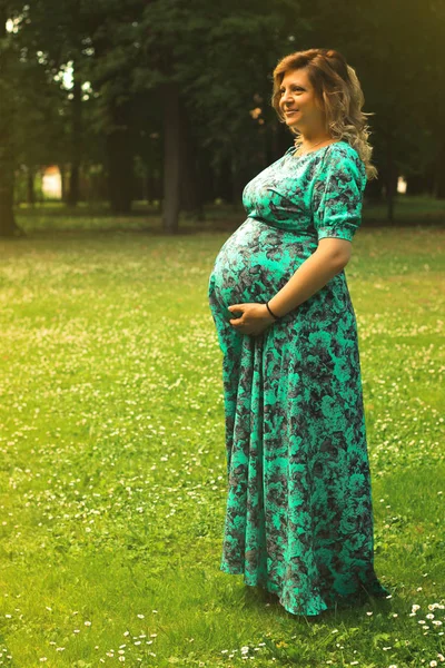 Uma jovem mulher grávida bonita segura as mãos em seu estômago em um vestido longo contra um fundo de árvores e gras verde no verão — Fotografia de Stock