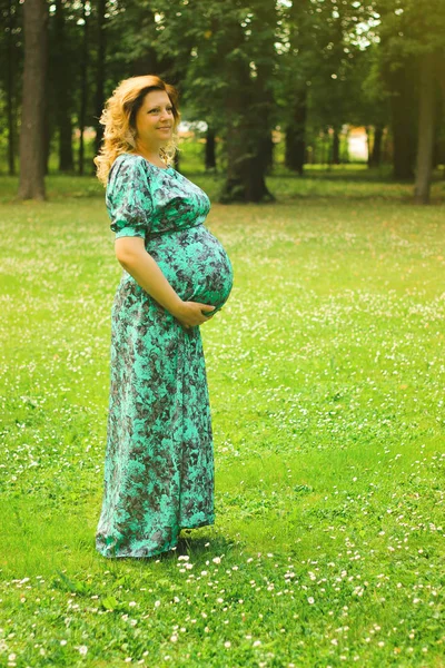 A young beautiful pregnant woman holds her hands on her stomach in a long dress against a background of trees and green gras  in summertime — Stock Photo, Image