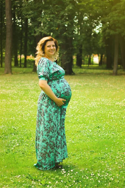 젊은 아름 다운 임신한 여자 보유 그녀의 손에 긴 복장에서 그녀의 속에 나무와 녹색 그의 배경에 대해 여름에 — 스톡 사진