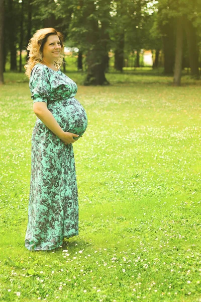 Uma jovem mulher grávida bonita segura as mãos em seu estômago em um vestido longo contra um fundo de árvores e gras verde no verão — Fotografia de Stock