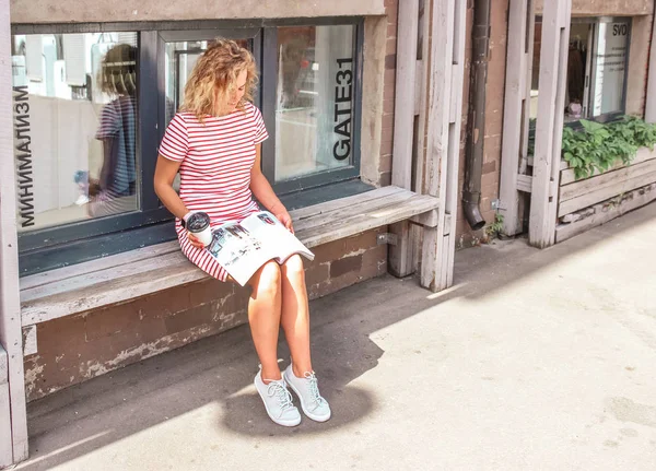 Jovem linda garota loira sentada no banco lendo notícias de moda na revista e bebendo café da manhã na cidade grande — Fotografia de Stock