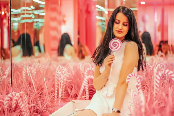 Young beautiful brunette girl on the soft pink background with multi-colored bright pink lollipop — Stock Photo, Image