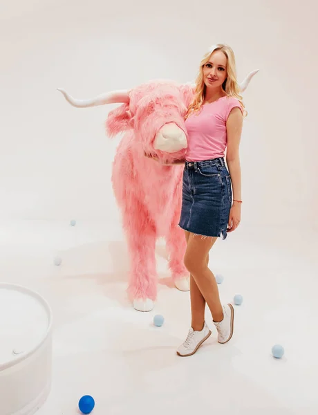 Jonge mooie blondie in witte jurk op een witte achtergrond met fantastisch landschap met een roze stier — Stockfoto