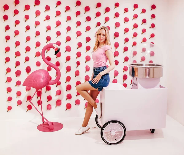 Mladá krásná blondýnka dívka pózuje na pozadí zeď s růžovou zmrzlinou a zařízení pro výrobu vaty sladké. Sladká dekorace na pozadí — Stock fotografie