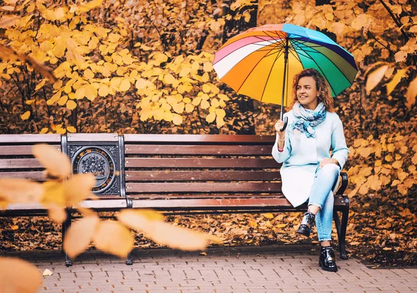 年轻美丽的女孩坐在长凳上的秋天公园与五颜六色的雨伞 — 图库照片
