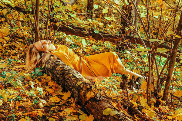 Young beautiful girl in a long yellow dress lying on the ground of the autumn park with fallen yellow leaves. Artistic processing.