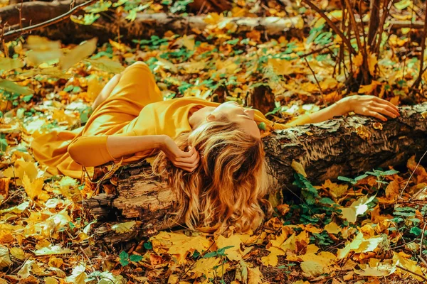 Young beautiful girl in a long yellow dress lying on the ground of the autumn park with fallen yellow leaves — Stock Photo, Image