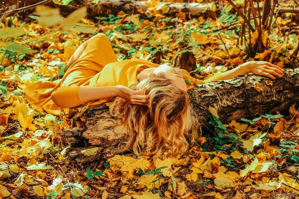 Giovane bella ragazza in un lungo abito giallo sdraiata sul terreno del parco autunnale con foglie gialle cadute — Foto Stock