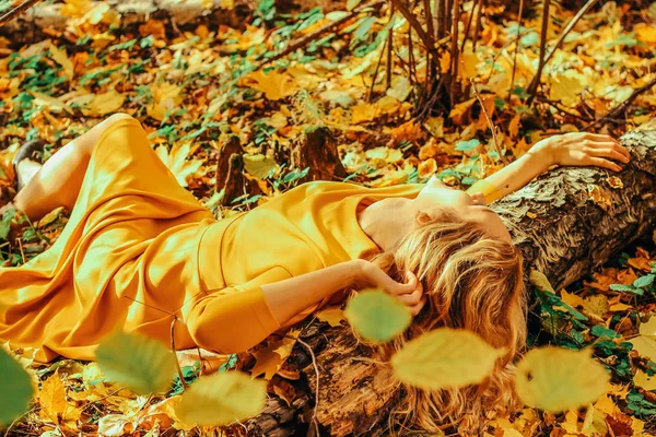 Young beautiful girl in a long yellow dress lying on the ground of the autumn park with fallen yellow leaves — Stock Photo, Image