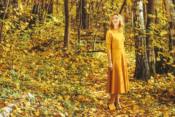 年轻美丽的女孩在黄色长礼服散步在秋季公园与落叶 — 图库照片
