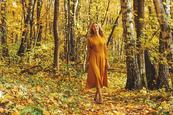 Joven hermosa chica en un vestido largo amarillo camina en el parque de otoño con hojas caídas —  Fotos de Stock