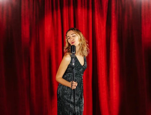 Mladá atraktivní dívka zpívá na pódiu s mikrofonem na pozadí červené závěsy — Stock fotografie