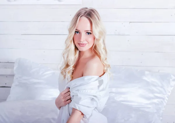Mladá sexy blondýna žena v bílé košili a sedí na bílé posteli v ateliéru — Stock fotografie