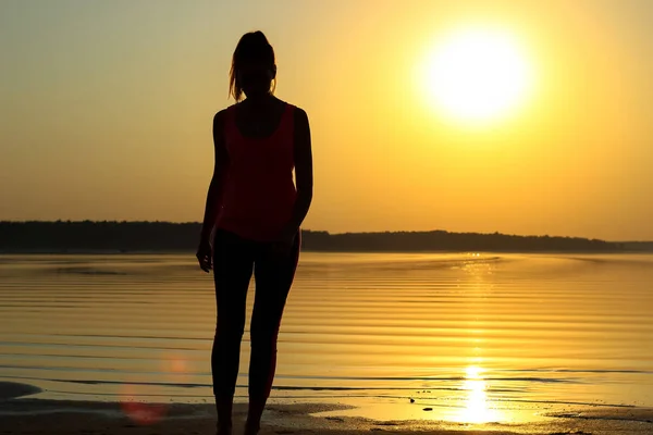Silhouette of a young beautiful girl walking along the seashore during orange sunset.