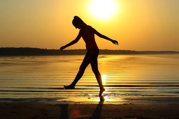 Silhouette of a young beautiful girl walking along the seashore during orange sunset.