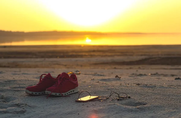 Sports set. Pink sneakers and a mobile phone with headphones close-up on a sandy beach by the sea during sunset.