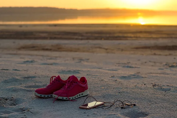 Sports set. Pink sneakers and a mobile phone with headphones close-up on a sandy beach by the sea during sunset.