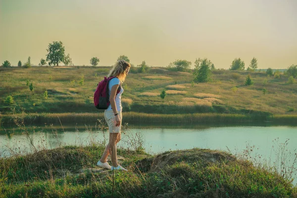 Young woman with pink backpack is standing on the blue chalk quarries background close up in sunset time