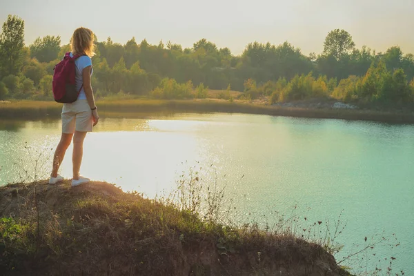 Young woman with pink backpack is standing on the blue chalk quarries background close up in sunset time