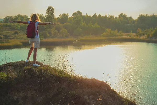 Young woman with pink backpack is standing on the blue chalk quarries background close up in sunset time