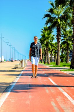 Genç çekici adam yaz aylarında palmiye ağaçları ile yaya yolu boyunca yürür, Karadeniz kıyısına yakın, Batum, Gürcistan
